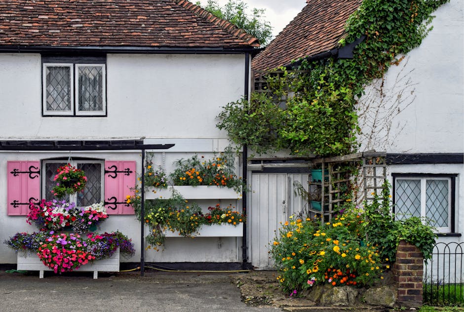 A small garden area in front of a white cottage with a single window featuring decorative white shutters and a small black wrought iron balcony. The garden is neatly organized with a central paved pat